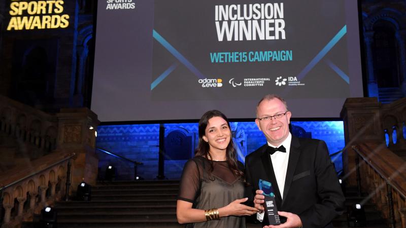 A man and a woman hold a trophy in front of a screen with the words &quot;Inclusion Winner WeThe15 Campaign&quot;