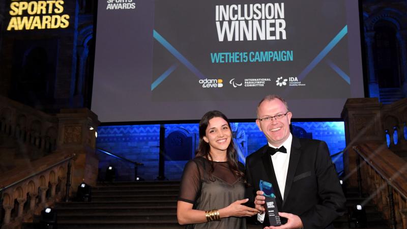 A man and a woman hold a trophy in front of a screen with the words "Inclusion Winner WeThe15 Campaign"