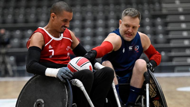 A male wheelchair rugby player wearing a red jersey carries the ball, while another player reaches for the ball.