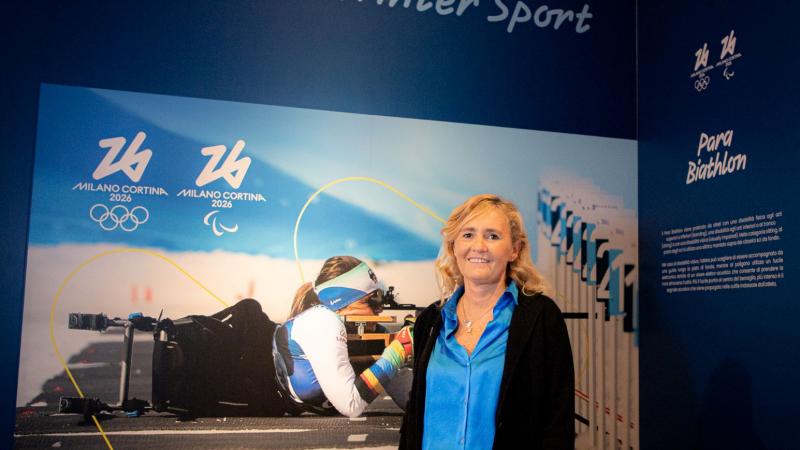 A woman in business attire poses in front of a poster featuring a Para biathlon athlete in action.