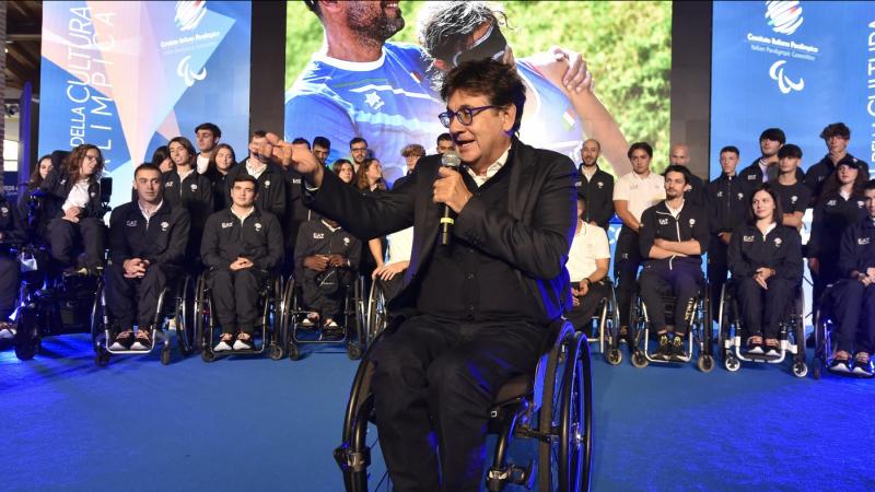 A man in a wheelchair speaks in a microphone on stage with a crowd of athletes behind him.