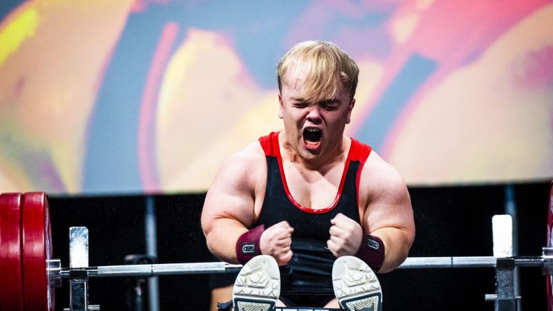 A male Para powerlifter of short stature screams and squeezes his fists in celebration after making a successful lift.