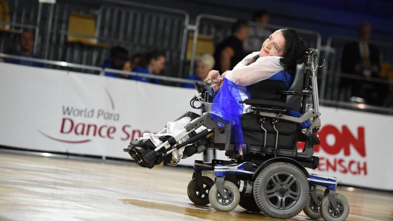 A female dancer in an electric wheelchair smiles and motions with her hands during a dance performance.