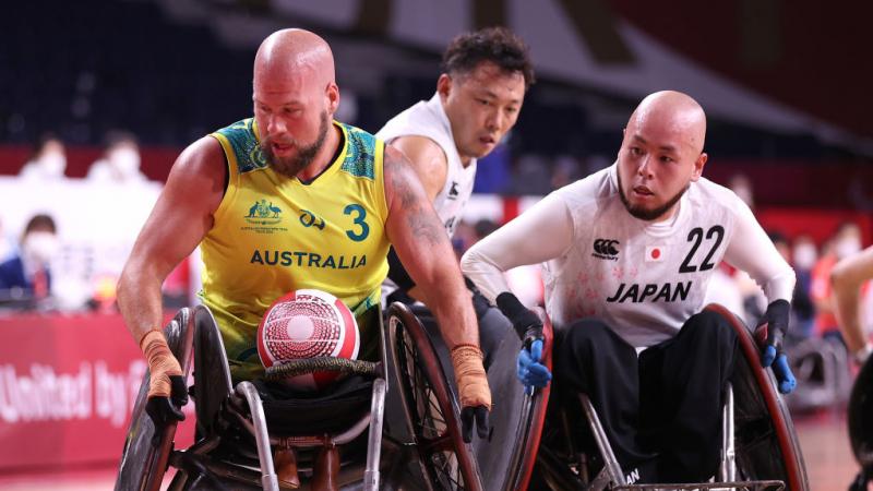 A male wheelchair rugby player in a yellow jersey carries the ball, chased by two players.