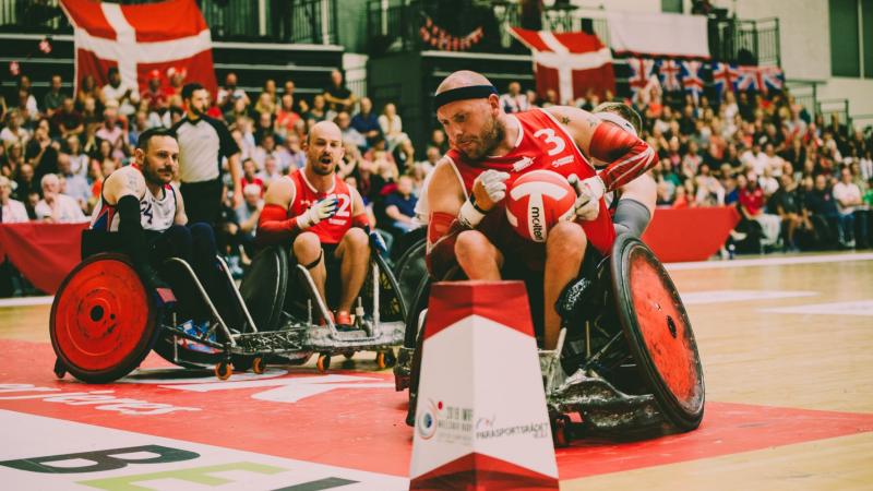 Three male athletes in wheelchairs, with one carrying a red and white ball, play at a venue packed with spectators.