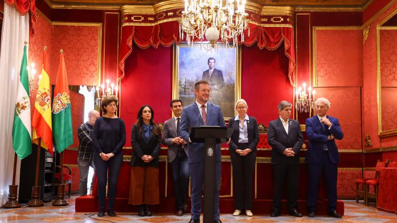 A man in a suit smiles while giving a speech at the podium in a lavishly decorated palace room.