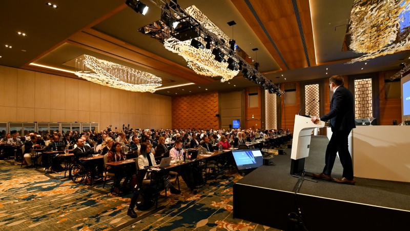 A man gives a speech from the stage as a room full of IPC member organisations looks on.