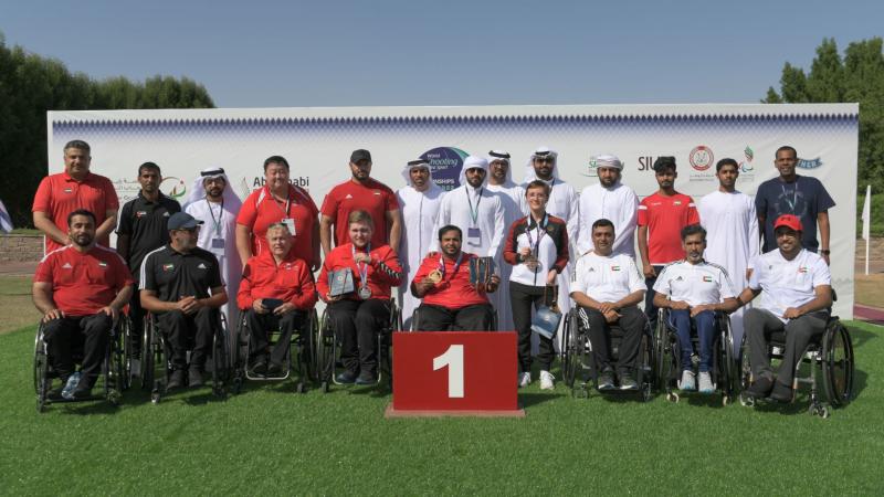 A group of nine men in wheelchairs posing in a podium with 14 people behind them