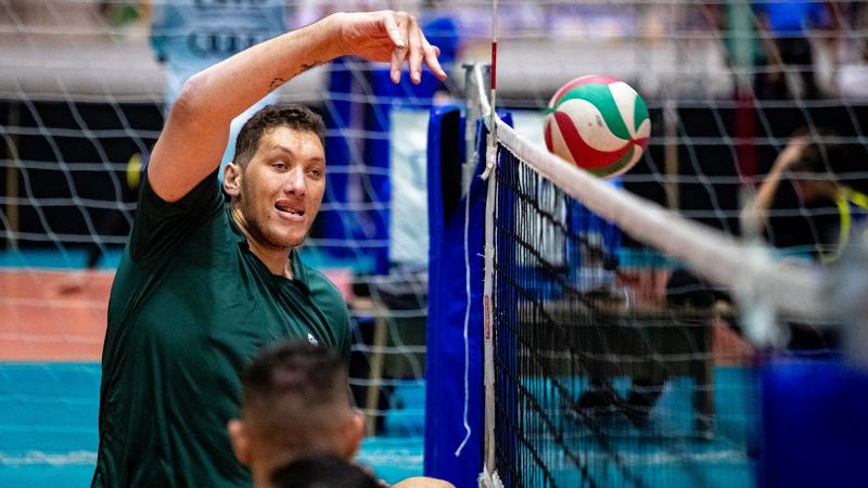 A male sitting volleyball player raises his right arm to hit the ball above the net