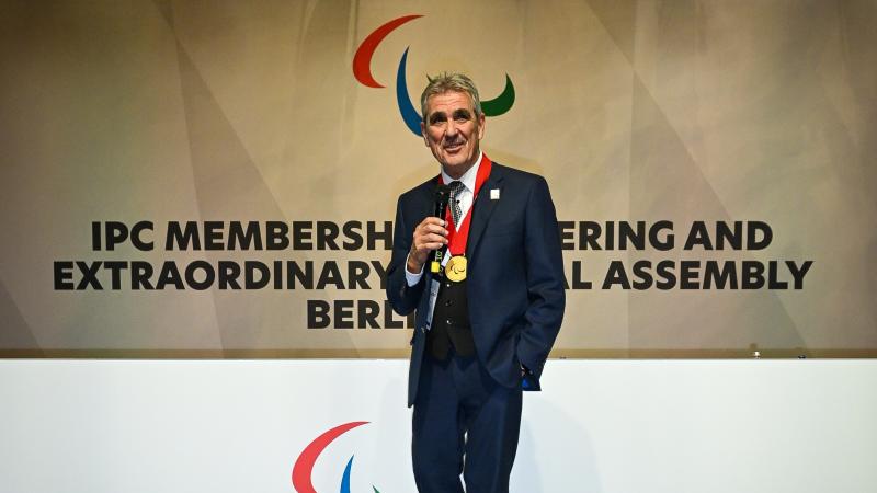 A man with a gold medal around his neck gives a speech from the stage with the Agitos logo behind him.