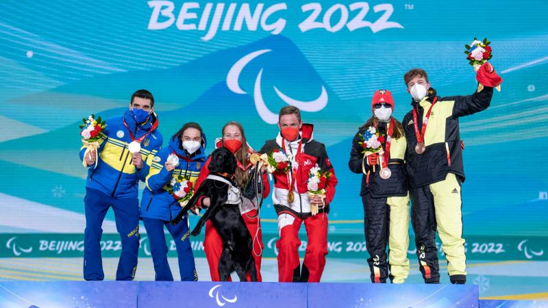 Six athletes pose for a photograph on the podium. A female athlete holds a gold medal with her left hand and her black guide dog with her right hand.