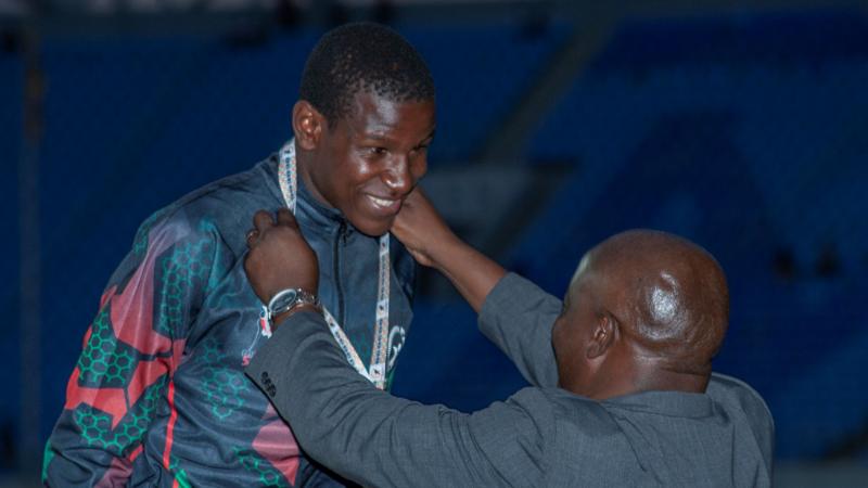 A male athlete in a national kit smiles as a medal presenter puts a medal around his neck.