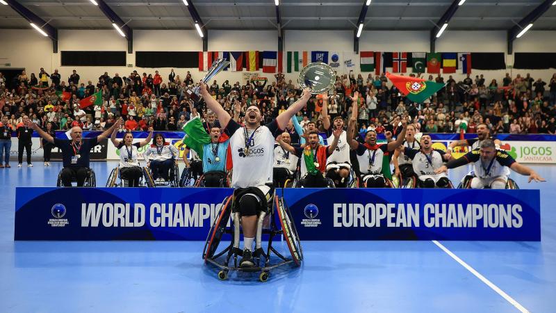 A male athlete in a wheelchair screams in celebration while holding up a trophy and dish, as a team cheers behind him next to the banner "world champions" and "European champions".