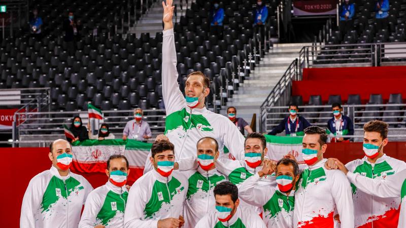 Male sitting volleyball athletes in national uniforms and masks featuring the Iranian flag pose for a photo as they wait for the medal ceremony at Tokyo 2020.