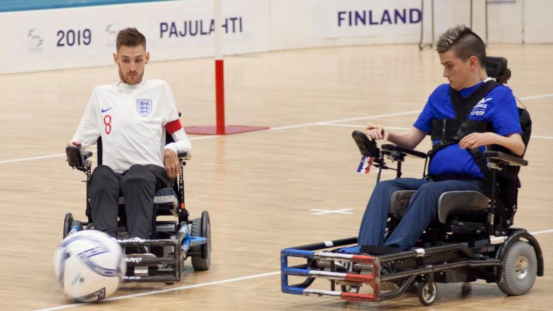 A male powerchair football athlete controls his wheelchair in front of the ball, while another player looks at the ball