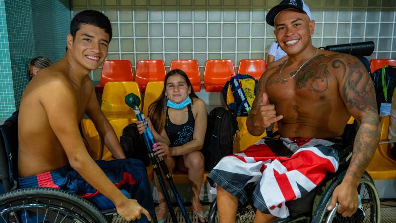Three swimmers, two males in wheelchairs and a female swimmer holding equipment for athletes with vision impairments, pose for a photo on the bleachers.