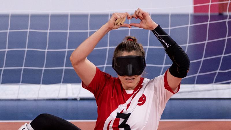 A female goalball player in an eye mask makes a heart gesture with her hands as she sits on the competition field.