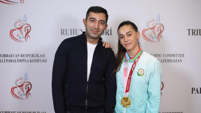 A man in and a woman, her right hand on his shoulder and a gold medal around her neck, pose in front of an official backdrop.