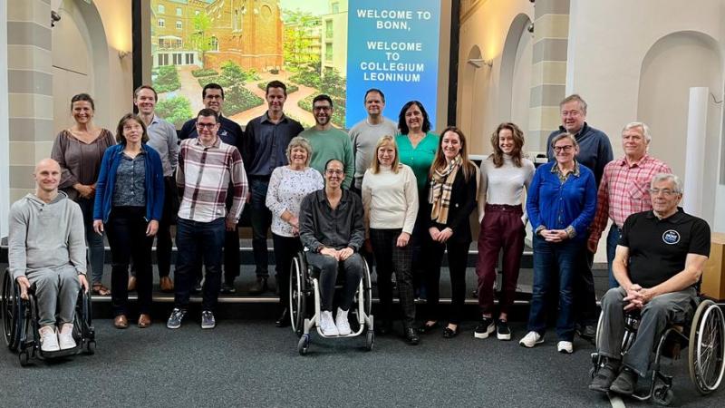 19 people pose for a photograph in front of a screen that says "Welcome to Bonn"