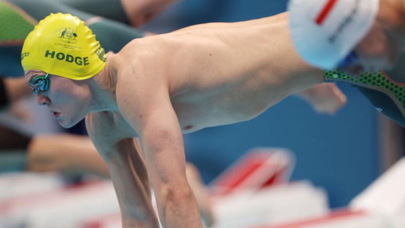 A male swimmer with a cap of Australia jumping in the water with another male swimmer in the foreground