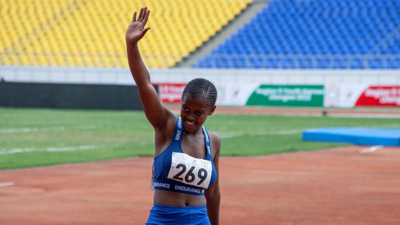 A female runner smiles and raises her right arm in a salute as she comes off the running track.