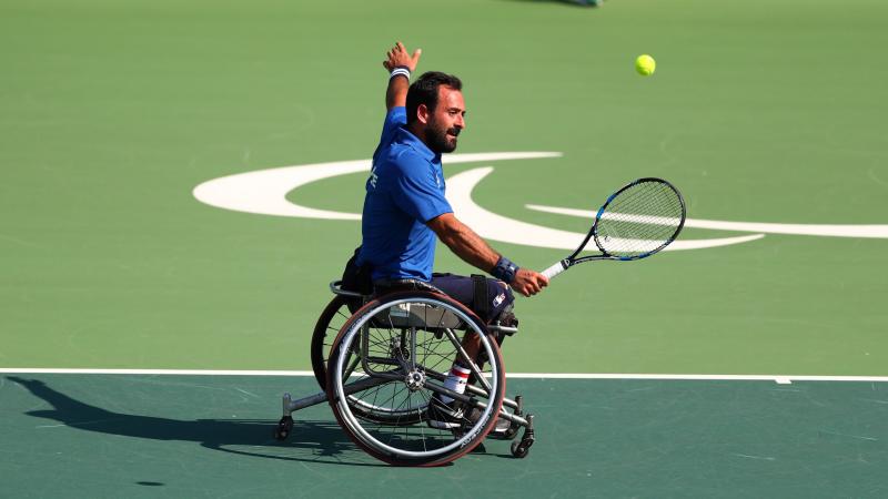 A male wheelchair tennis player makes a serve on a hard green court with his left arm stretched out wide.