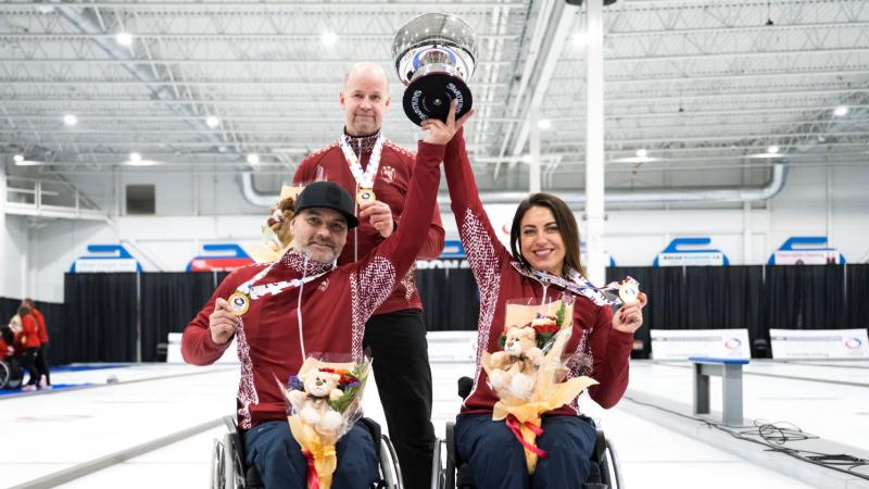 A female wheelchair curler and a male wheelchair curler lift a trophy together, while showing a gold medal