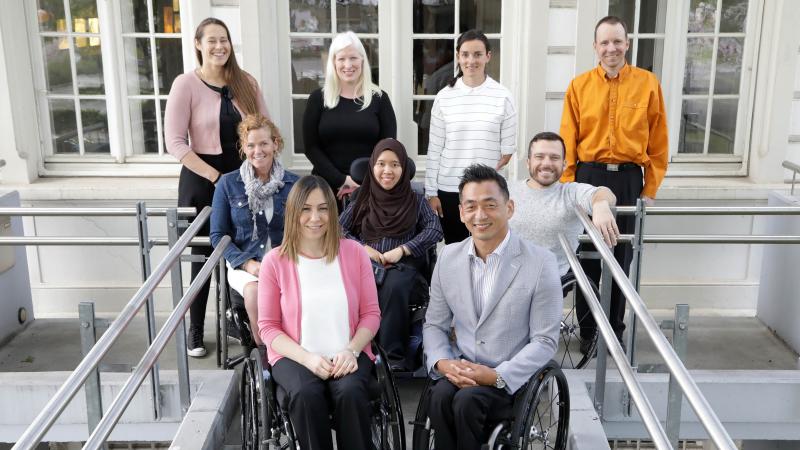 Nine athletes, five in wheelchairs at the front and second rows, and four standing in the back pose for a photo in front of the IPC headquarters.