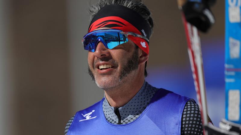 A close-up of a male cross-country skier in sunglasses and competition bib, holding skis in one hand. 