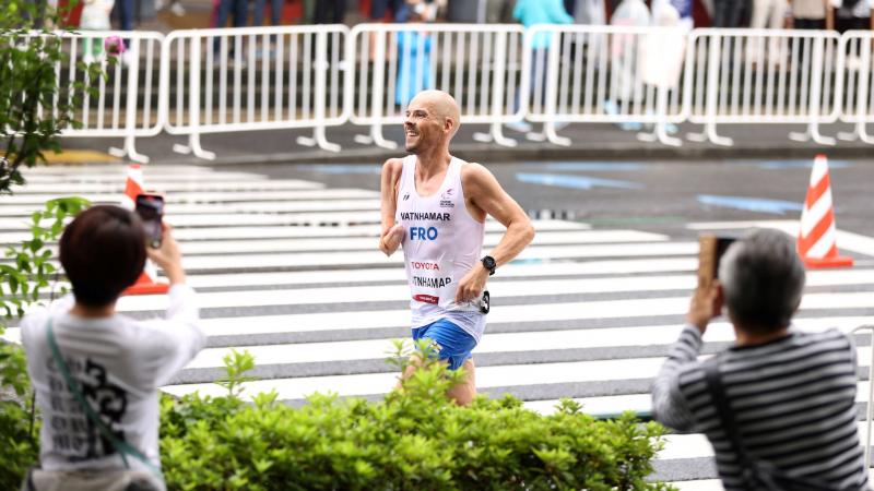 An athlete who is missing his right arm below the elbow smiles as he runs on the street during a marathon as onlookers take photos of him.