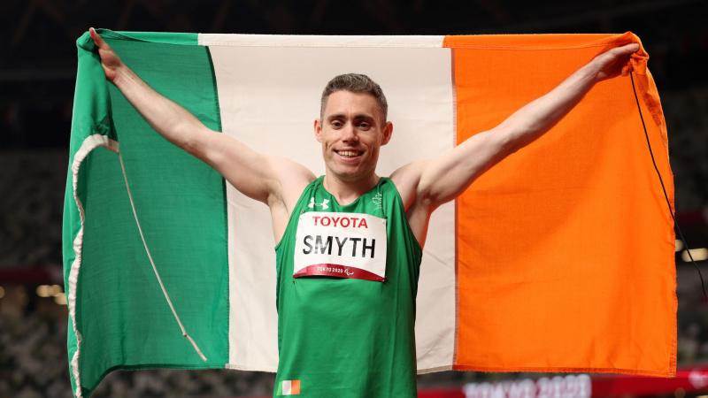 A male athlete holds up an Irish flag behind him as he celebrates a victory in the athletics stadium at Tokyo 2020.