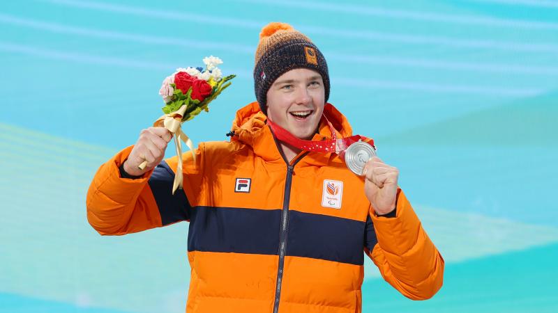 A smiling male athlete holds up a silver medal and bouquet of flowers during a medal ceremony. 