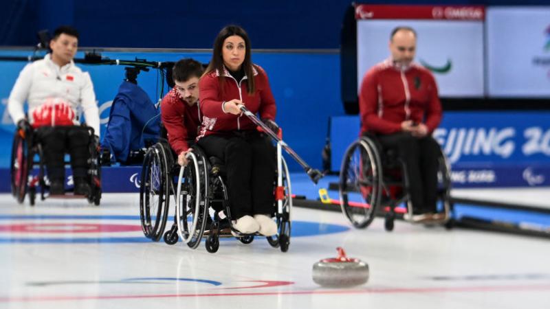 A female wheelchair curler delivers a stone with a stick, while a male athlete holds the wheels of her wheelchair.