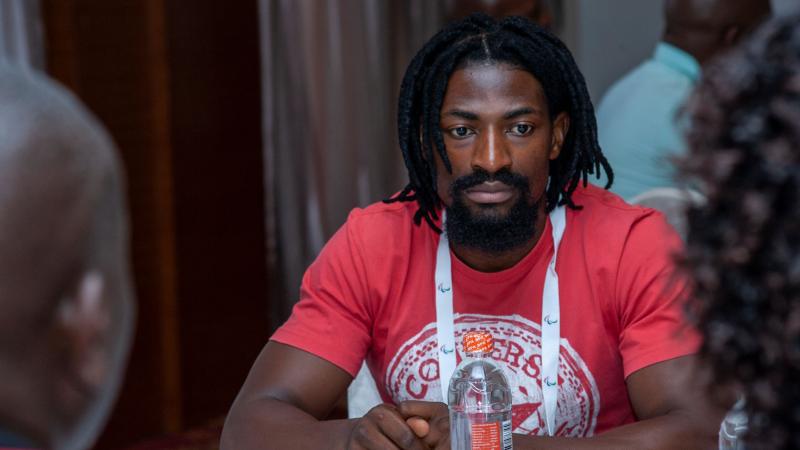 A man in a red t-shirt looks on as he sits at a table with his hands folded in front of him.