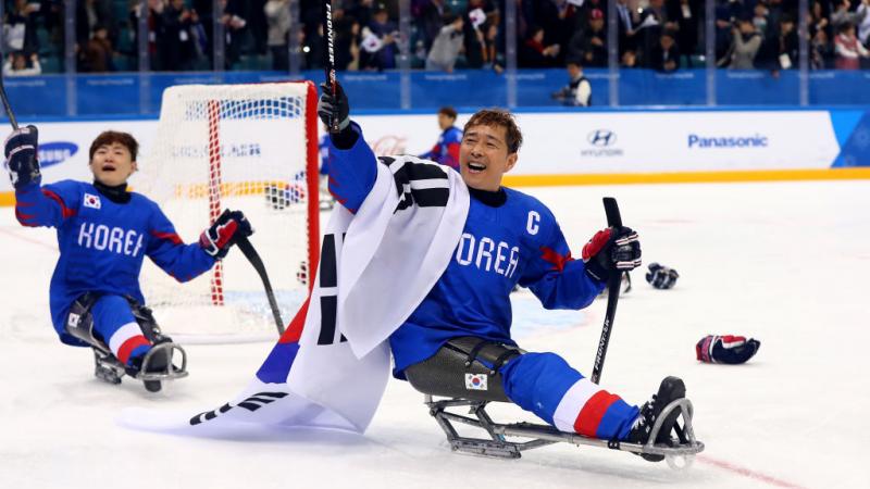 A male Para ice hockey player holds up a sledge hockey stick with his right hand and carries the Republic of Korea flag.