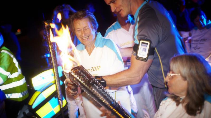 A woman in a wheelchair passes the Paralympic Flame through a torch to another woman, also holding a torch.