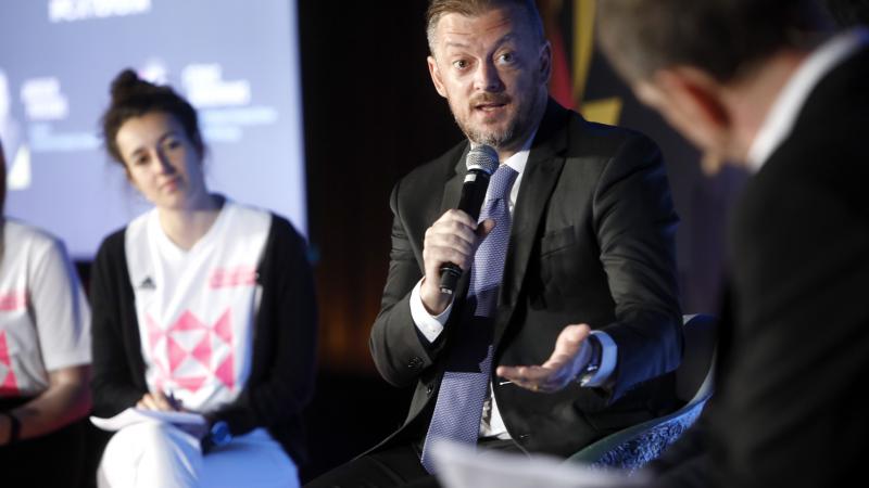 A man in a suit speaks into a microphone during a discussion session at Global Sport Week in Paris. 