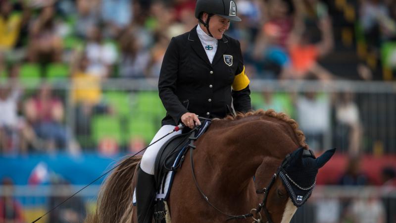 A female Para equestrian rider smiles as she rides a horse during competition.