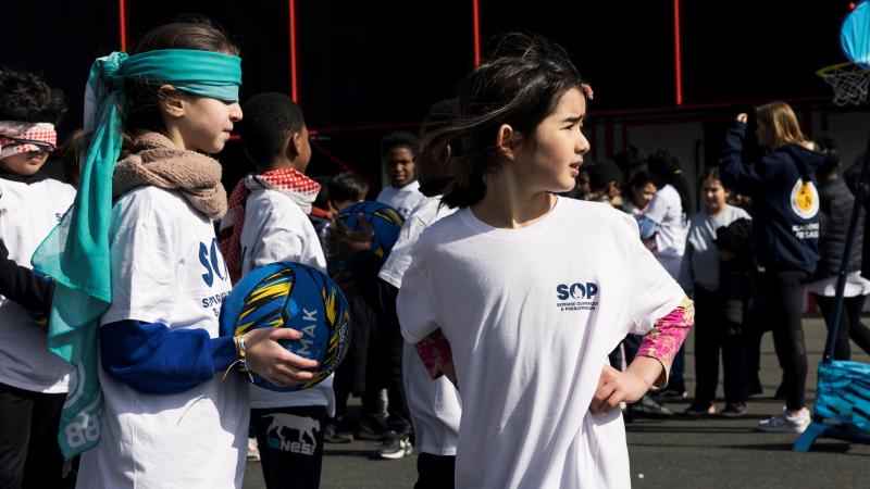 Two young girls, one with a blindfold, holding a ball, and the other looking back, participate in an event during Olympic and Paralympic Week.