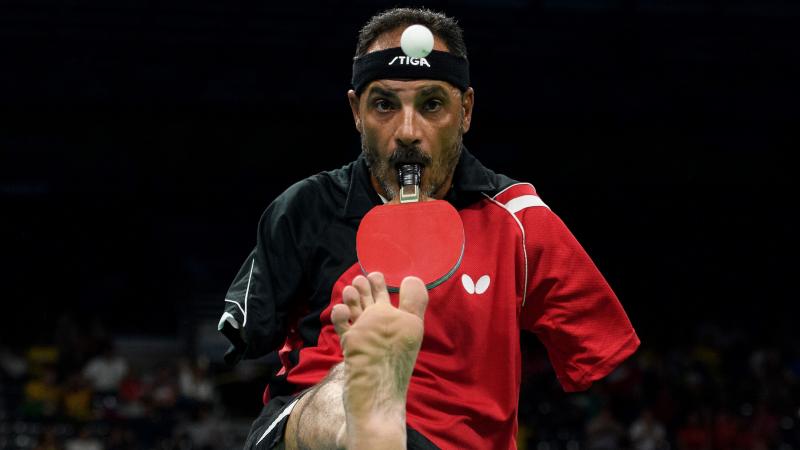 A male Para table tennis player holds a racquet with his mouth and lifts the ball with his right foot.