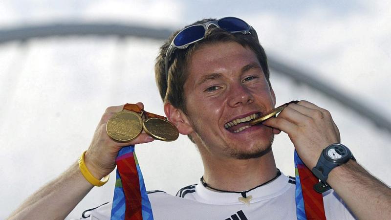 A male athlete poses for a photo by biting a gold medal and raises two other gold medals in his right hand.