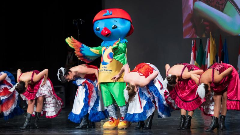 The mascot of the Virtus Global Games, which is a blue bird wearing a red cap, on a stage with seven women who are bowing.