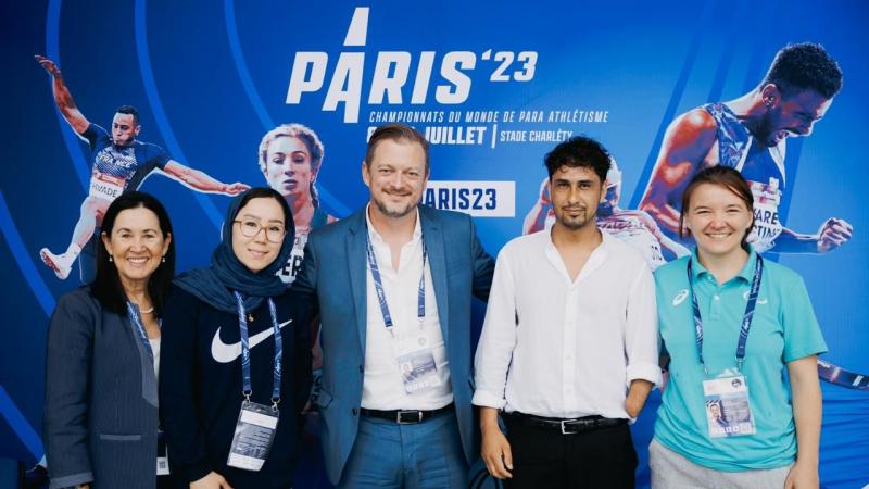 Five people pose for a group photograph in front of a Paris 23 Para Athletics World Championships banner