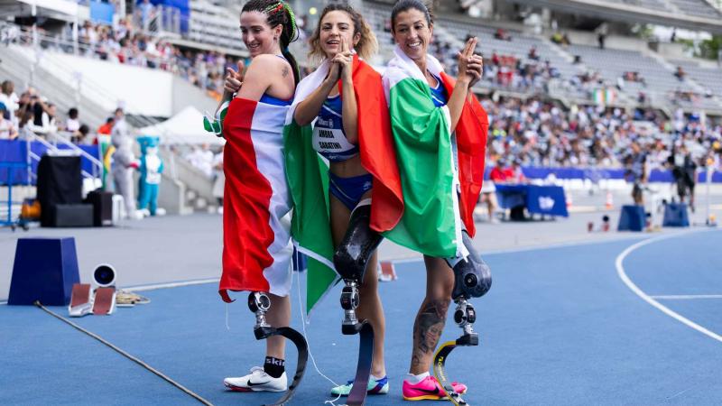 Three female athletes with prosthetic legs posing for a picture with the flag of Italy