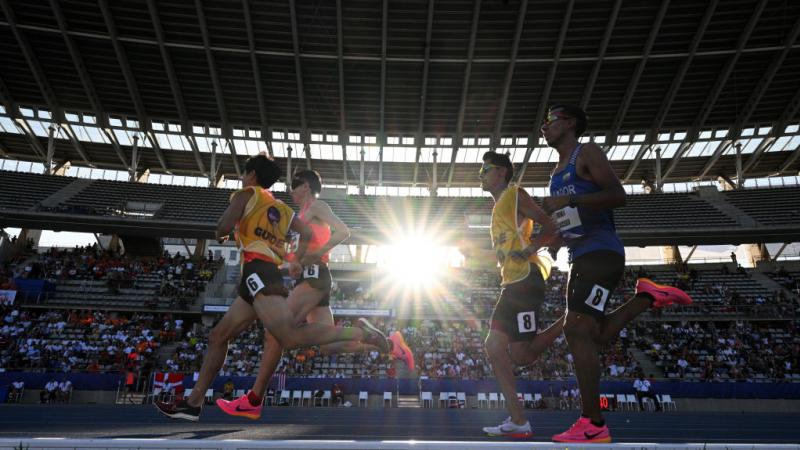 two vision impaired runners and their guides running on the track in front of spectators 