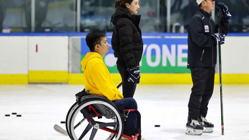 A man in a wheelchair next to a standing woman and a man on an ice rink