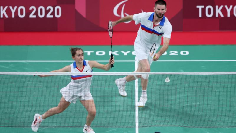 Photo shows a mixed doubles badminton match. A female athlete runs to a shuttle, in front of a male athlete.