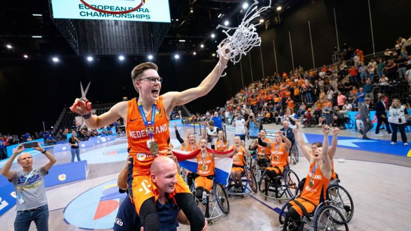 A female wheelchair basketball player cuts a basketball net, her teammates celebrate