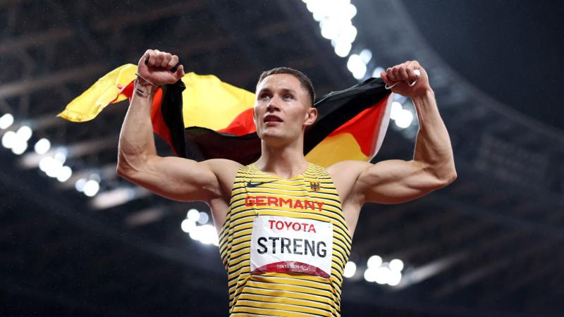 A male athlete holds the German flag with both hands at a stadium.