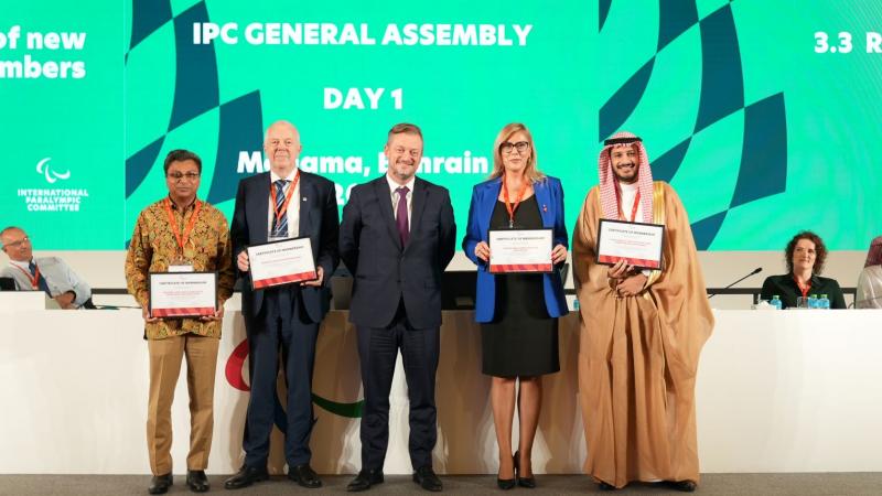 Five people pose for a group photo in front of a green screen that says "IPC General Assembly".